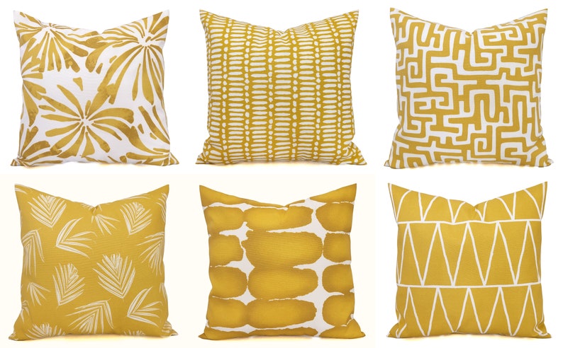 May include: Six decorative throw pillows with various geometric and botanical patterns in mustard yellow and white. Designs include abstract shapes, linear patterns, and leaf motifs. The pillows are square and appear to be made of fabric.