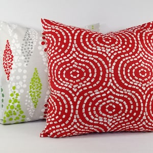 May include: Two square throw pillows. The front pillow is bright red with a white dashed circular pattern. The back pillow is light gray with a pattern of vertical diamond shapes composed of small red, white, and lime green dots.