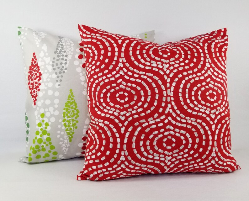 May include: Two square throw pillows. The front pillow is bright red with a white dashed circular pattern. The back pillow is light gray with a pattern of vertical diamond shapes composed of small red, white, and lime green dots.