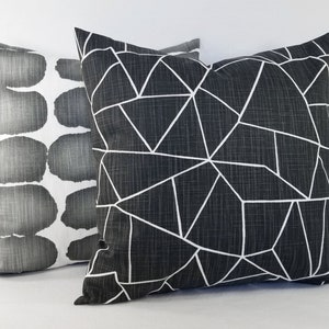 May include: Two square scatter cushions. The front cushion is dark charcoal grey with a white geometric line pattern. The back cushion is white with a repeating pattern of dark grey, rounded, horizontal shapes. Both cushions have a textured fabric finish.