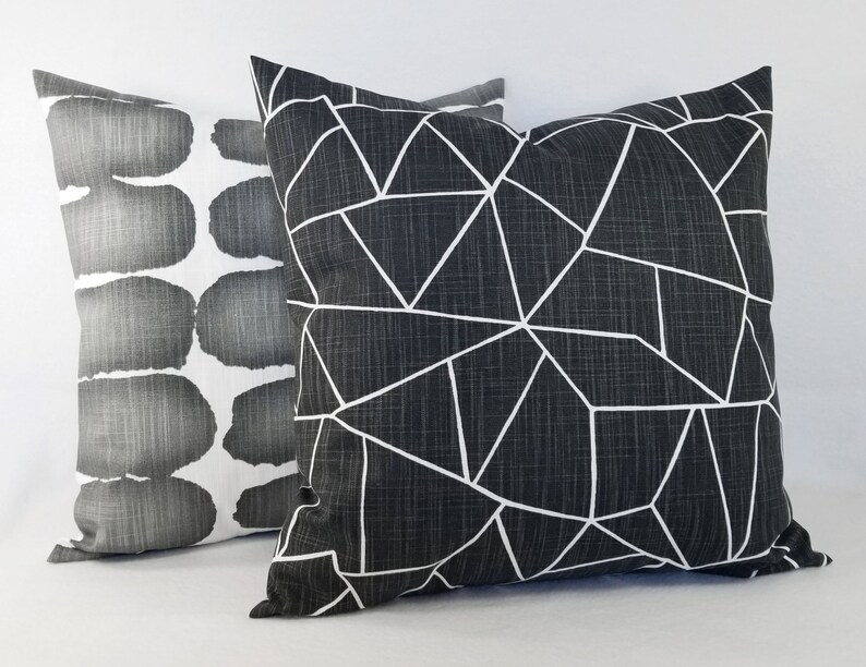 May include: Two square scatter cushions. The front cushion is dark charcoal grey with a white geometric line pattern. The back cushion is white with a repeating pattern of dark grey, rounded, horizontal shapes. Both cushions have a textured fabric finish.