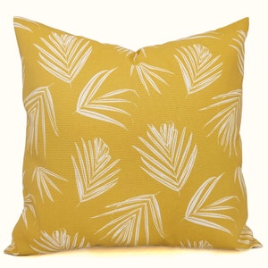 May include: Square decorative pillow with a yellow background and a white palm leaf pattern. The pillow is a square shape and appears to be made of a textured fabric. The design is a tropical theme.
