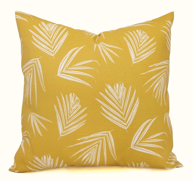 May include: Square decorative pillow with a yellow background and a white palm leaf pattern. The pillow is a square shape and appears to be made of a textured fabric. The design is a tropical theme.