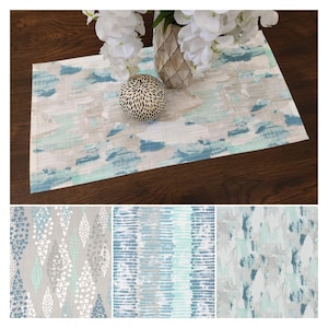 May include: A rectangular table runner with an abstract design in shades of blue, gray, and white. The runner is placed on a dark wooden table, with a vase of white flowers and a decorative sphere on top. Three additional fabric samples are shown below.
