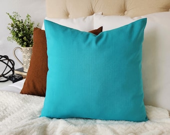 May include: A turquoise blue throw pillow on a white faux fur bedspread. The pillow is square and has a simple design.