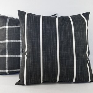 May include: Two square throw cushions featuring a dark charcoal grey fabric with a visible woven texture. One cushion displays a white vertical striped pattern, while the other cushion features a white grid or plaid pattern across the dark surface.
