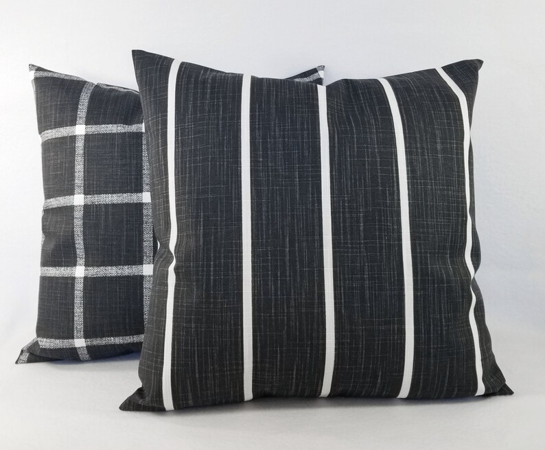 May include: Two square throw cushions featuring a dark charcoal grey fabric with a visible woven texture. One cushion displays a white vertical striped pattern, while the other cushion features a white grid or plaid pattern across the dark surface.