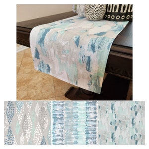 May include: A set of three table runners with abstract patterns in shades of blue, green, and gray. The runners are made of a soft, linen-like fabric and feature a variety of brushstroke designs.