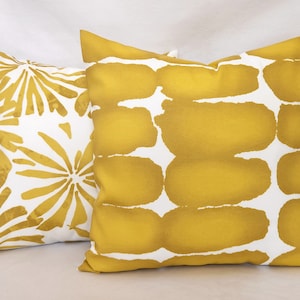 May include: Two decorative throw pillows. One pillow features a white background with a gold floral pattern. The other pillow has a white background with a gold abstract design. The pillows are square and made of fabric.