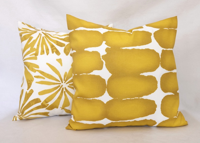 May include: Two decorative throw pillows. One pillow features a white background with a gold floral pattern. The other pillow has a white background with a gold abstract design. The pillows are square and made of fabric.