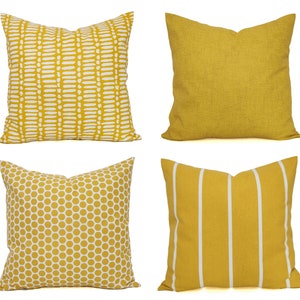 May include: A set of four square throw pillows in shades of mustard yellow and white. The collection includes one pillow with a white dash pattern, one solid mustard yellow pillow, one with a white polka dot print, and one with vertical white stripes.
