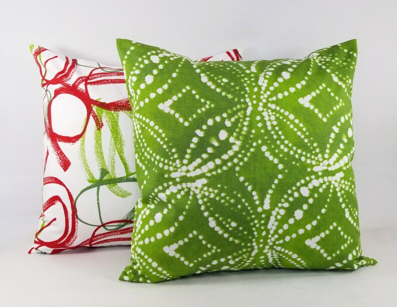 May include: Two square throw pillows. The front pillow features a vibrant lime green fabric with a white dotted geometric pattern. The back pillow is white with a bold red and green abstract circular and linear print design on the fabric.