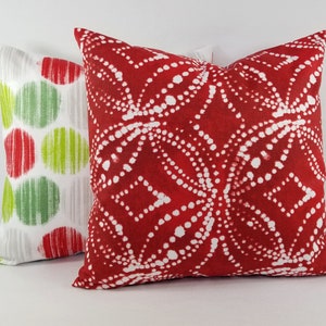 May include: Two square throw pillows. The front pillow is deep red with a white dotted geometric pattern. The back pillow is white with a repeating pattern of red and lime green circles, each featuring vertical line textures within the shapes.