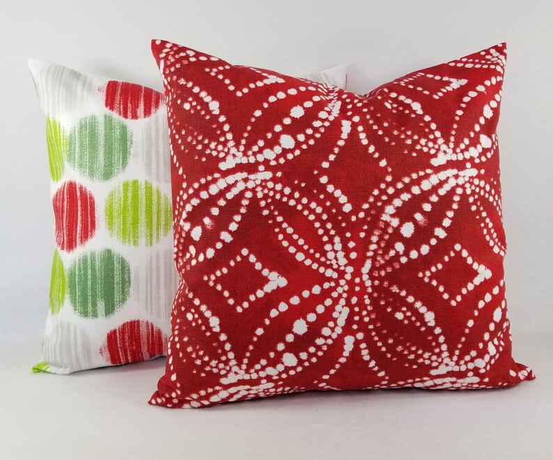 May include: Two square throw pillows. The front pillow is deep red with a white dotted geometric pattern. The back pillow is white with a repeating pattern of red and lime green circles, each featuring vertical line textures within the shapes.