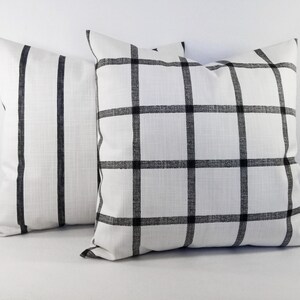 May include: Two square throw cushions featuring a white fabric base with black patterns. One cushion displays a grid of black lines forming a windowpane check, while the other cushion features vertical black stripes. Both cushions have a textured finish.