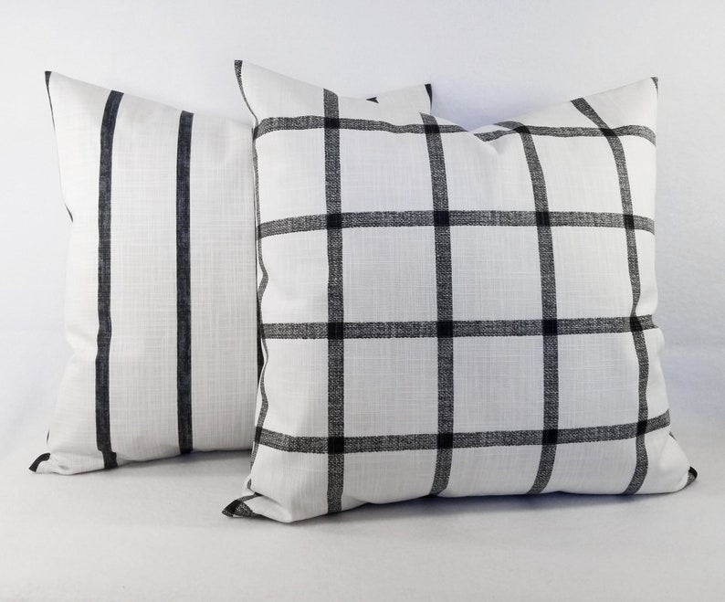 May include: Two square throw cushions featuring a white fabric base with black patterns. One cushion displays a grid of black lines forming a windowpane check, while the other cushion features vertical black stripes. Both cushions have a textured finish.
