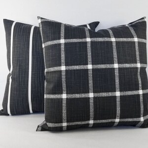 May include: Two square scatter cushions featuring a dark charcoal grey fabric with a visible woven texture. One cushion displays a white grid pattern, while the other features two vertical white stripes running down the centre of the dark surface.
