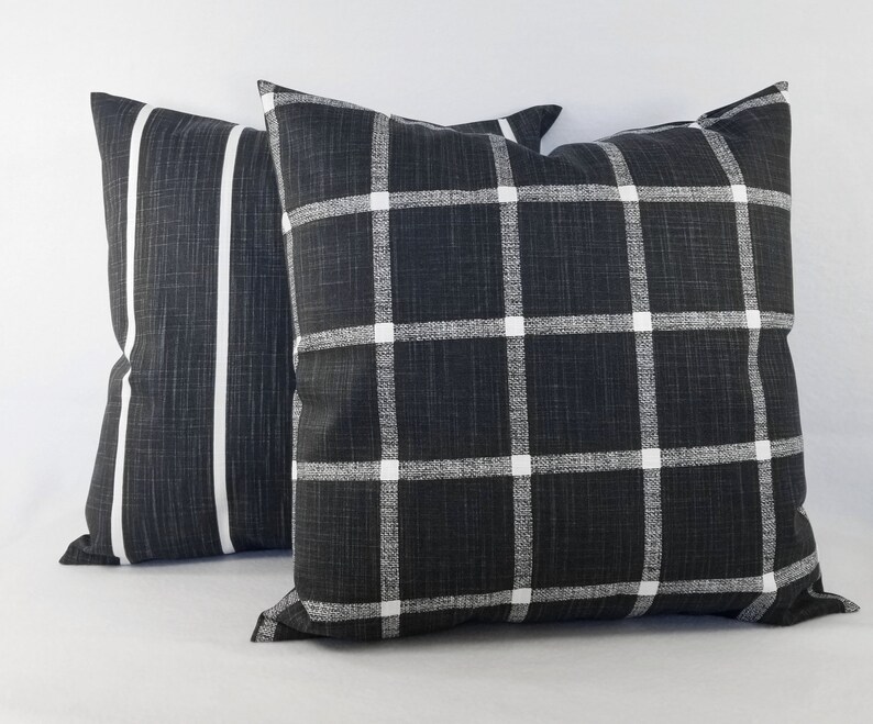 May include: Two square scatter cushions featuring a dark charcoal grey fabric with a visible woven texture. One cushion displays a white grid pattern, while the other features two vertical white stripes running down the centre of the dark surface.