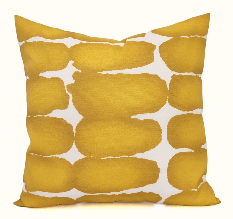 May include: Square decorative pillow with a mustard yellow and white abstract pattern. The pillow features a repeating design of large, irregular shapes in a warm yellow hue against a white background. The pillow is a square shape.