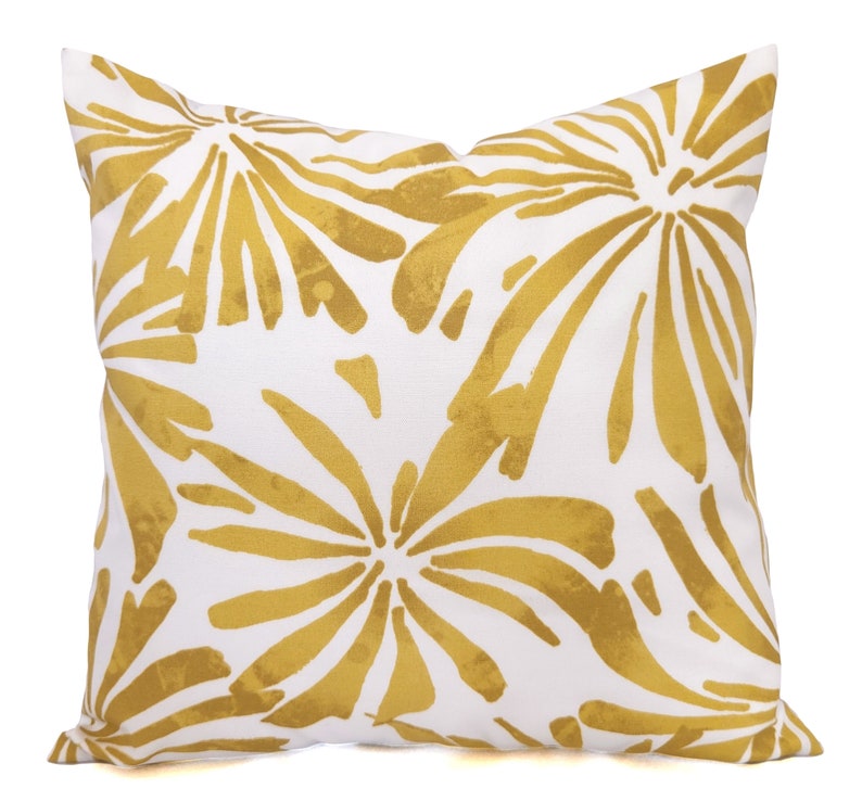 May include: Square white throw pillow with a gold floral pattern. The design features large, stylized flowers in a golden hue against a white background. The pillow is a decorative accent for a sofa or bed.