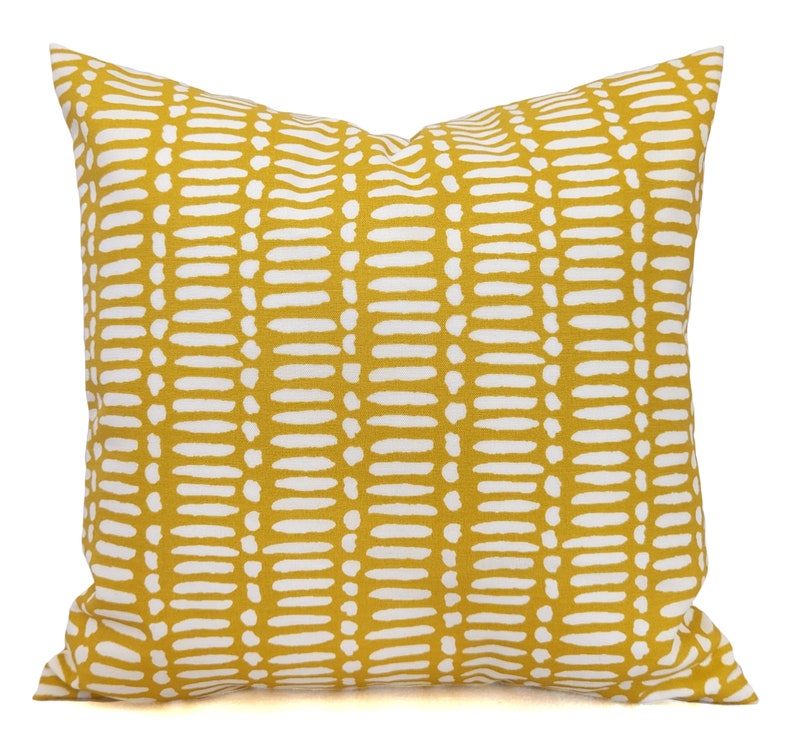May include: Square decorative pillow with a mustard yellow background and a repeating pattern of white dashes. The pillow has a soft, textured appearance and is likely made of fabric. The design is modern and would complement various home decor styles.