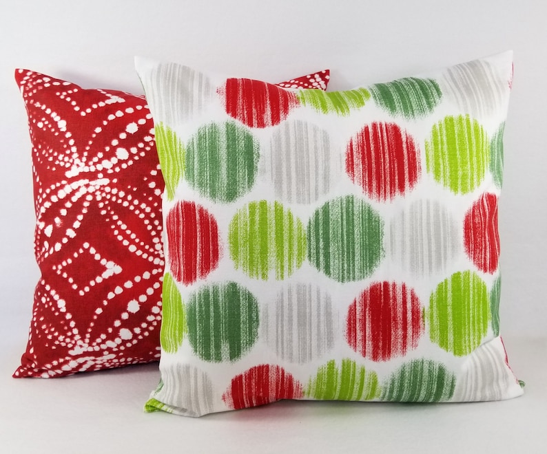 May include: Two square throw pillows. The front pillow features a white background with a repeating pattern of red, lime green, and light gray circles. The back pillow is red with a white dotted geometric pattern arranged in a diamond motif.