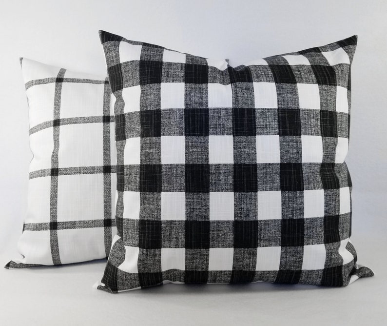 May include: Two square throw cushions featuring a black and white pattern. The front cushion displays a dense black and white buffalo check pattern, while the cushion positioned behind it features a larger, sparse black and white grid pattern.