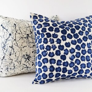 Puede incluir: Dos almohadas decorativas con fondo blanco y patrones azules. Una almohada tiene un dise&ntilde;o de pintura salpicada, mientras que la otra tiene un patr&oacute;n de lunares azules y blancos.