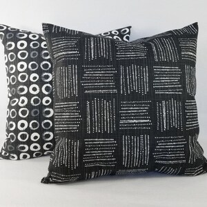 May include: Two square throw cushions featuring black fabric with white printed patterns. The front cushion displays a grid of alternating horizontal and vertical dashed lines, while the back cushion features a repeating pattern of white circles.