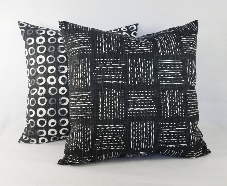 May include: Two square throw cushions featuring black fabric with white printed patterns. The front cushion displays a grid of alternating horizontal and vertical dashed lines, while the back cushion features a repeating pattern of white circles.