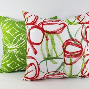 May include: Two square throw pillows. The front pillow is white with a red and green scribbled circular pattern. The back pillow is solid green with a white dotted geometric pattern. Both pillows feature a soft fabric texture and square shape.
