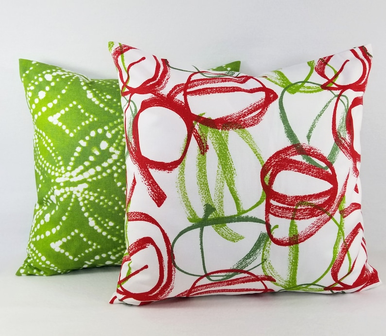 May include: Two square throw pillows. The front pillow is white with a red and green scribbled circular pattern. The back pillow is solid green with a white dotted geometric pattern. Both pillows feature a soft fabric texture and square shape.