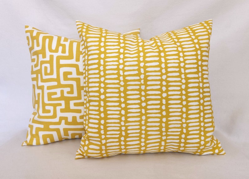 May include: Two decorative throw pillows with mustard yellow fabric and white patterns. One pillow features a maze-like design, while the other has a dotted pattern. The pillows are square and appear to be made of a soft fabric.