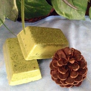 May include: Two bars of handmade soap with a green and yellow color. The soap has a speckled appearance. The soap is sitting on a blue and white fabric with a brown pine cone.