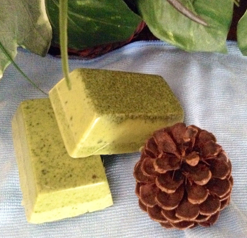 May include: Two bars of handmade soap with a green and yellow color. The soap has a speckled appearance. The soap is sitting on a blue and white fabric with a brown pine cone.