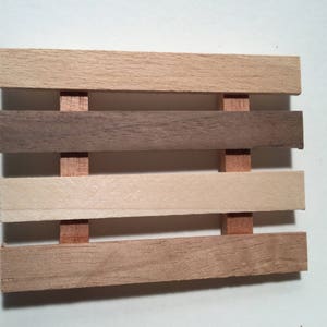 May include: A wooden soap dish with four slats, each a different shade of wood. The top slat is light brown, the second is dark brown, the third is light beige, and the bottom slat is a medium brown.