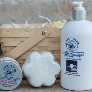 Puede incluir: Una cesta de regalo que contiene una botella blanca de loción de leche de cabra y miel, una bomba de baño blanca con forma de flor y una lata blanca de manteca corporal sedosa de leche de cabra. Los tres productos tienen una etiqueta con el texto "A Circle of Old Goats at The Spa" y el texto "Ylang Ylang & Lavender for a Restful Night's Sleep".