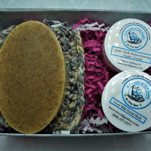 May include: A gift box filled with pink and white shredded paper contains a crocheted washcloth, a bar of goat milk soap, and two small jars of goat milk skin care products. The jars are labeled "A Couple of Old Goats at The Spa" and contain goat milk rejuvenating night cream and goat milk facial mask.