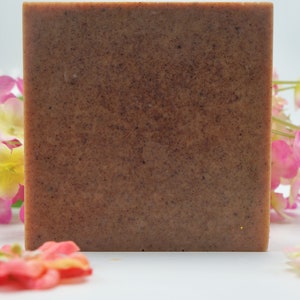 May include: A square, brown soap bar with a textured surface. The soap is surrounded by pink, yellow, and orange flowers on a white background. The soap appears to be handmade.