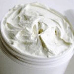 May include: A white, creamy body butter in a white plastic jar. The butter is thick and smooth, and the jar is open, revealing the product inside.