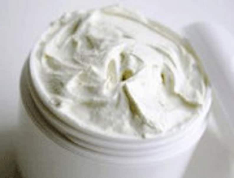 May include: A white, creamy body butter in a white plastic jar. The butter is thick and smooth, and the jar is open, revealing the product inside.