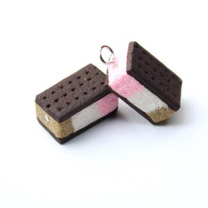 May include: Two rectangular ice cream sandwich charms featuring dark brown textured wafers with a dotted pattern. The filling consists of layered pink and white sections. One charm includes a small silver metal jump ring attached to the top edge.