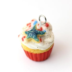 May include: A miniature cupcake charm featuring a yellow cake base in a red fluted liner, topped with white frosting, small red and blue sprinkles, gold glitter, and a central dark blue glittery starfish, finished with a silver metal jump ring.