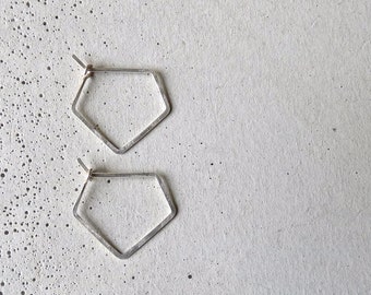 May include: A pair of silver-toned, geometric hoop earrings. Each earring has a five-sided, pentagon shape with a textured surface. The earrings are displayed on a speckled, light gray surface.