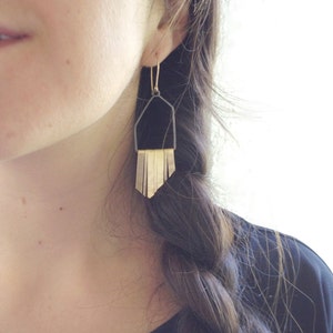 Könnte beinhalten: Ein Paar goldene und schwarze geometrische Ohrringe mit Fransen. Die Ohrringe hängen am Ohrläppchen und haben oben eine schwarze geometrische Form.