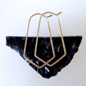 May include: A pair of gold-toned geometric hoop earrings. The earrings are made of thin metal and have a slightly irregular shape.