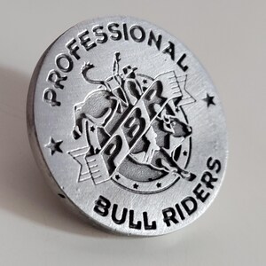 May include: A silver-colored round metal emblem with the words "PROFESSIONAL BULL RIDERS" engraved around the edge. The center features a detailed image of a bull rider on a bucking bull, with the letters "PBR" prominently displayed.