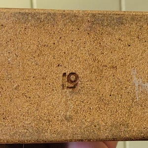 May include: Close-up of a rectangular, light brown object with a textured surface. The number "19" is stamped in the center. There are some white marks on the right side of the object.