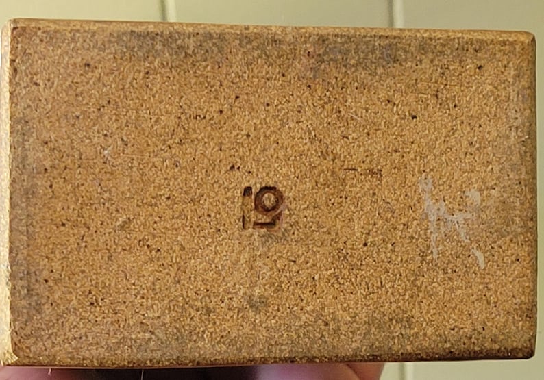 May include: Close-up of a rectangular, light brown object with a textured surface. The number "19" is stamped in the center. There are some white marks on the right side of the object.