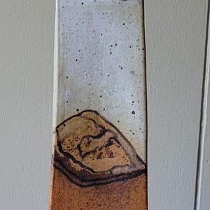 May include: Tall, rectangular ceramic vase with a narrow neck. The upper portion is off-white with black speckles, transitioning to a brown, textured lower section. The vase has a unique, organic shape and is likely handmade.